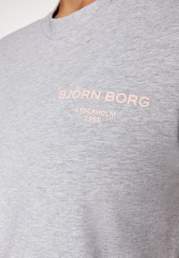 Lys grå skjorte med lys pink tekst "BJÖRN BORG STOCKHOLM 1956" på den øverste brystet, vist i nærbillede.