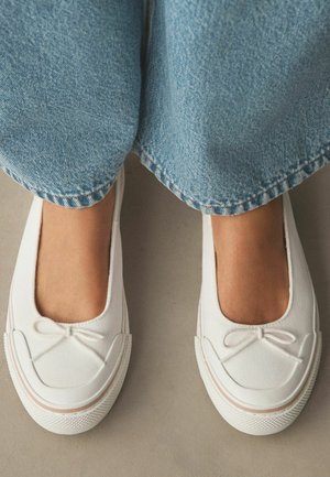 Stopy w białych butach wsuwanych z małymi kokardkami, ubrany w jasnoniebieskie dżinsy na neutralnym tle podłogi.