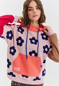 Mikina s kapucňou z ružovej látky s námorníckymi kvetinovými vzormi, oranžovou kapsou, modrými šnúrkami a rukávmi v kombinácii ružovej a hnedej farby.