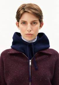 Jonge persoon met kort bruin haar, die een donkerblauwe gebreide capuchon over een lichte coltrui en een bordeauxrode spikkelvest met ritssluiting draagt.