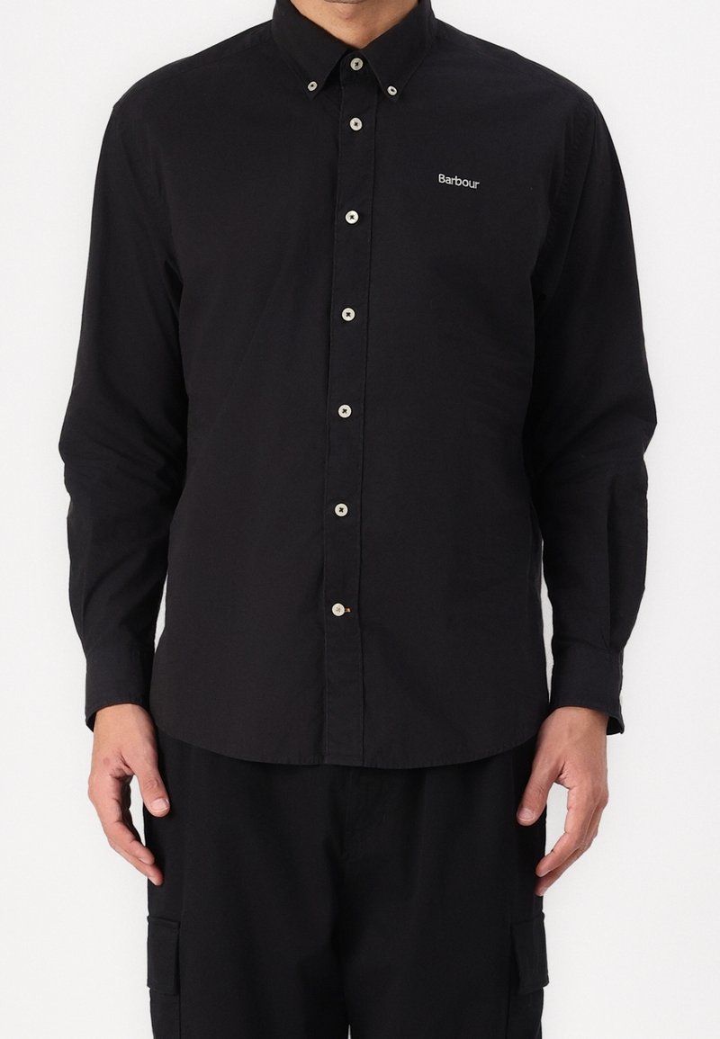 Homme portant une chemise noire à manches longues avec des boutons blancs et le logo Barbour sur le haut de la poitrine.