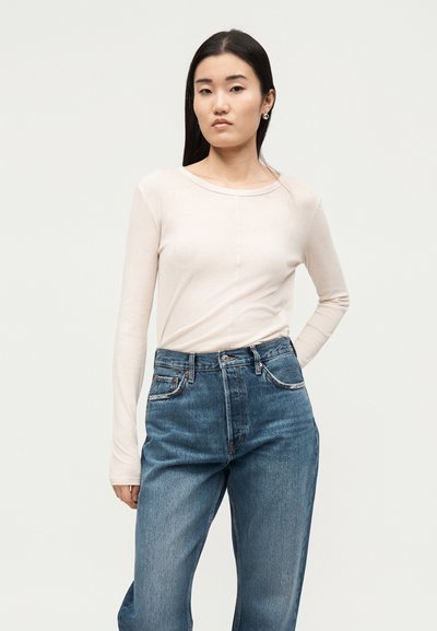 Νεαρή γυναίκα με ανοιχτό μπεζ μακρυμάνικη μπλούζα και ψηλόμεσο μπλε τζιν, στέκεται μπροστά σε απλό ανοιχτό φόντο με το ένα χέρι στη μέση.