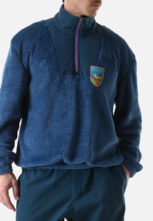 Homme portant un pull polaire bleu texturé avec demi-fermeture éclair et écusson coloré Millet, associé à un pantalon bleu foncé, main dans la poche.