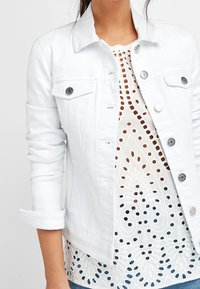 White denim jacket with metallic buttons and pockets, layered over a white patterned top featuring cut-out designs and intricate detailing.