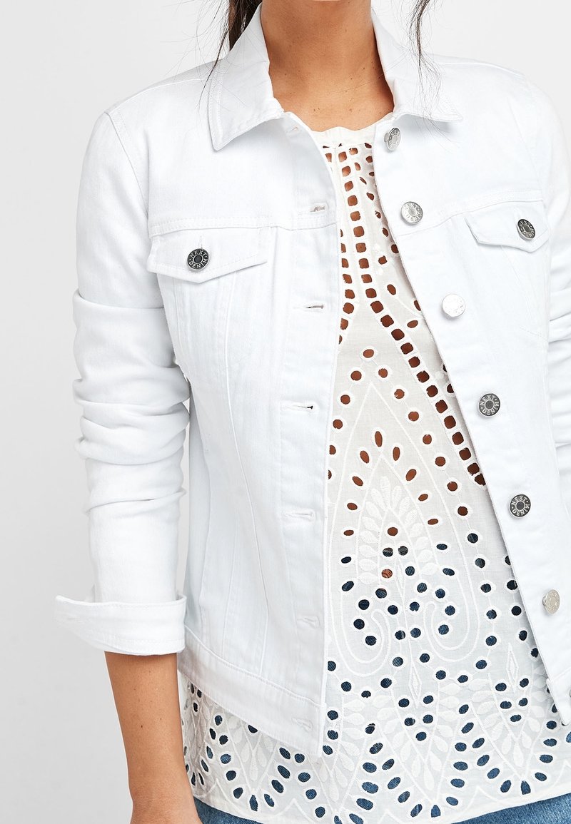 White denim jacket with metallic buttons and pockets, layered over a white patterned top featuring cut-out designs and intricate detailing.