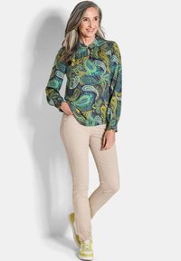 Langemouwen blouse met een paisleypatroon in groen, geel en blauw; gecombineerd met beige skinny pants en sneakers met gele accenten.