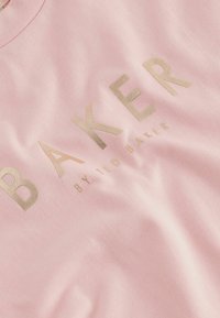 Rozā kokvilnas T-krekls ar apaļu kakla izgriezumu, uz kura ir zelta folijas drukājums "BAKER" un zem tā minimalistiskā dizainā norādīts "by TED BAKER".