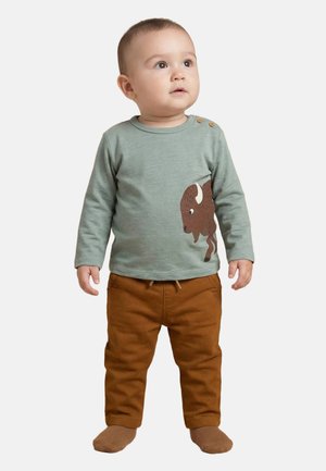 Tout-petit en chemise à manches longues verte avec un motif de buffle et pantalon marron, debout et regardant légèrement vers le haut à droite.