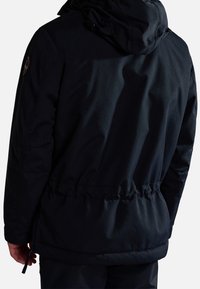 Vue de dos d'une personne portant une veste noire à capuche avec une taille cintrée et un patch logo sur la manche gauche.