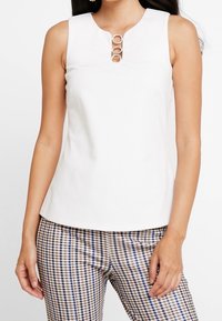 Top bianco senza maniche con scollatura rotonda, dotato di tre accenti metallici circolari sul davanti. Abbinato a pantaloni a quadri.