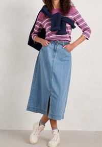 Jasnoniebieska dżinsowa spódnica midi z otworem z przodu, w połączeniu z fioletową koszulą w paski z długim rękawem i granatowym swetrem z dzianiny. Casuowe trampki.