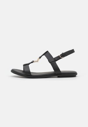 Sort flad sandal med tynde remme, guldfarvet spænde på den forreste rem og justerbar spænde rundt om anklen på hvid baggrund.