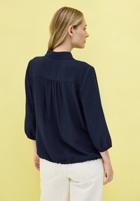Blusa leggera di colore blu navy con colletto, maniche a tre quarti e dettaglio arricciato sul retro, abbinata a pantaloni chiari.