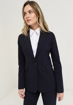 Kobieta o blond włosach, ubrana w dopasowaną czarną marynarkę i białą koszulę z kołnierzykiem, stojąca na jednolitym jasnym tle.
