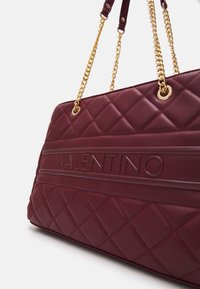 Burgundy quiltad handväska med guldkedjor och prägling av "VALENTINO"-logotyp. Har en strukturerad form och en slät lädertextur.
