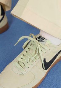 Lyse beige Nike-sneakers med sort swoosh, parret med cremede bukser, hvilende på en blå overflade. Snørebåndene er løst bundet.