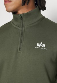 Felpa zip-up verde oliva con colletto alto, caratterizzata da un logo bianco sul petto e un tessuto liscio e testurizzato.