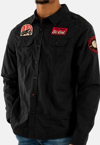 Camisa negra de botones hecha de algodón con dos bolsillos frontales. Presenta parches bordados en rojo y blanco, incluyendo "Von Dutch" y "So. Cal."