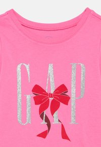 Rozā audums ar vārdu "GAP" lieliem sudraba mirdzošiem burtiem, rotāts ar sarkanu loku, kas ir piesiets ap burtu "A".