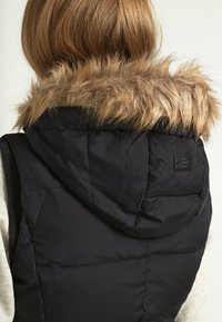 Osoba w czarnej puchowej kamizelce z kapturem wykończonym futrem, pokazująca widok z tyłu i włosy w kolorze jasnobrązowym sięgające ramion.