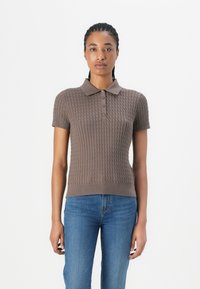 Frau mit geflochtenem Haar, die ein kurzärmliges braunes Strick-Polohemd und eine blaue Jeans trägt, steht vor einem schlichten weißen Hintergrund.