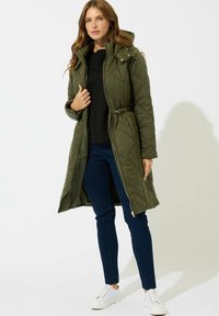 Mujer vestida con un abrigo largo acolchado de color verde oliva sobre una blusa negra, jeans azul oscuro y zapatillas blancas, de pie frente a un fondo liso.