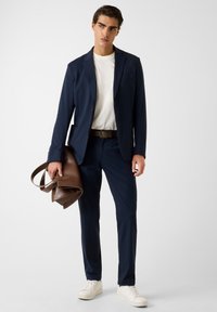 Marineblauer Anzug mit schmaler Passform, kombiniert mit einem weißen T-Shirt, einem braunen Ledergürtel und einer braunen Tasche. Das Model trägt weiße Sneaker.