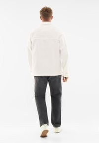 White denim jacket with large back pocket and button detail, paired with dark gray jeans and white sneakers, viewed from the back.