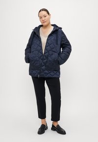 Marineblå quiltejakke med hette, glidelås og sidelommer. Båret over en beige topp og paret med svarte bukser og loafers.