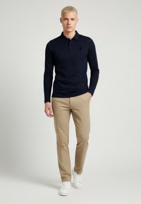 Polo shirt a maniche lunghe blu navy con colletto, abbinato a pantaloni beige texturizzati e sneakers bianche. Presenta un logo discreto sul petto.