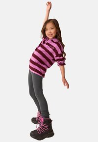 Jong meisje dat naar achter leunt met één arm omhoog, gekleed in een roze en bordeaux gestreepte trui, donkere leggings en zwarte laarzen met roze veters.