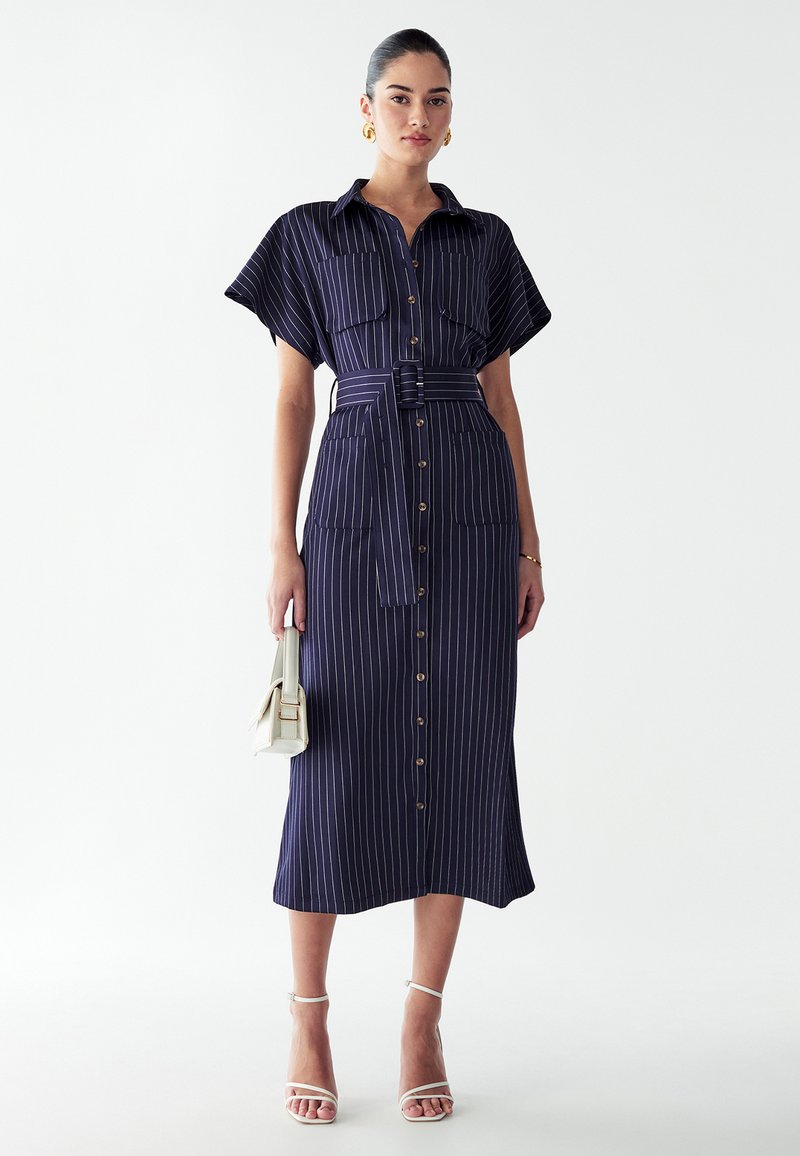 Jeune femme portant une robe midi boutonnée à rayures bleu marine avec manches courtes, taille ceinturée, talons blancs, et tenant un petit sac à main blanc.