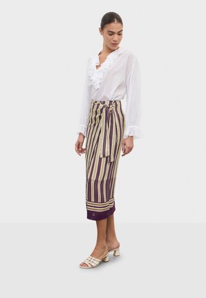 Žena v bielej blúzke s volánmi, prúžkovanej balerínovej midi sukni so šnúrkou a bielych sandáloch na podpätku stojaca pred jednoduchým pozadím.