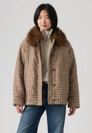 Donna che indossa un cappotto marrone pied-de-poule con un ampio colletto in pelliccia, un maglione beige con zip e jeans blu su uno sfondo uniforme.