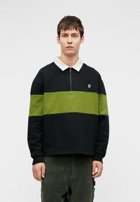 Jaunas vyras su trumpais rudais plaukais, vilkintis juodai ir žaliai juostuotą megztinį su balta apykakle ir tamsiai žaliomis kordūrinėmis kelnėmis, stovi prieš paprastą foną.