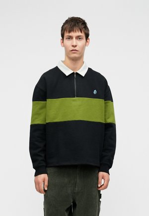 Fiatal férfi rövid barna hajjal fekete és zöld csíkos pulóvert visel fehér gallérral és sötétzöld kordbársony nadrággal, egy sima háttér előtt áll.