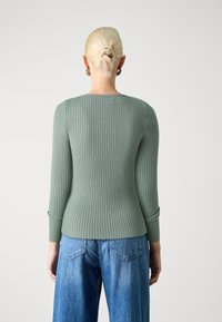 Jasnozielony sweter w prążki z długimi rękawami, dopasowany krój, okrągły dekolt i podwinięte mankiety, zestawiony z niebieskimi spodniami typu wide-leg.