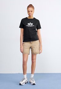 T-shirt nera in cotone con logo bianco "Alpha Industries", pantaloni corti in denim beige e sneakers grigie con accenti. Posizione in piedi su sfondo blu.