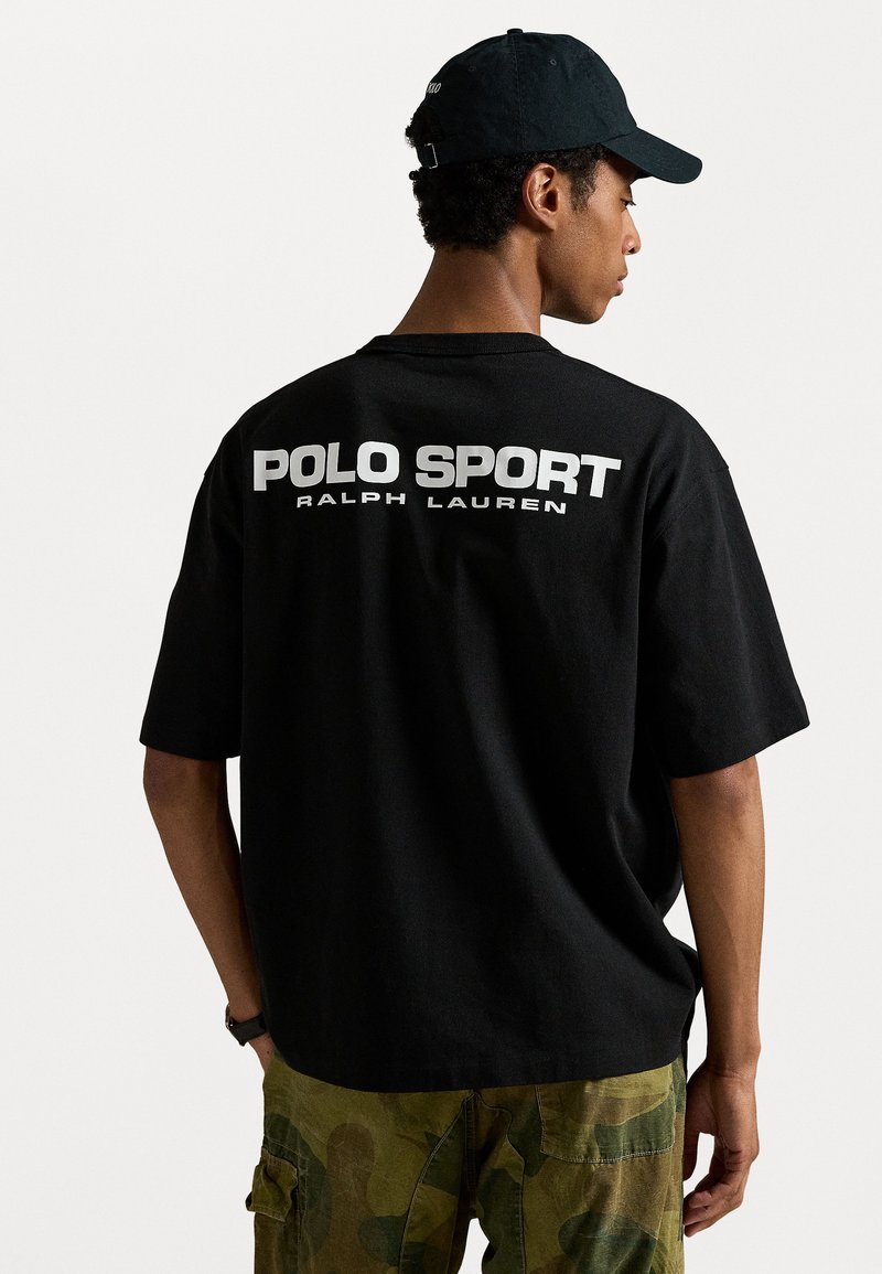 Zwarte katoenen t-shirt met "POLO SPORT" en "RALPH LAUREN" in het wit op de achterkant gedrukt. Gedragen met camouflage cargobroek en een zwarte pet.