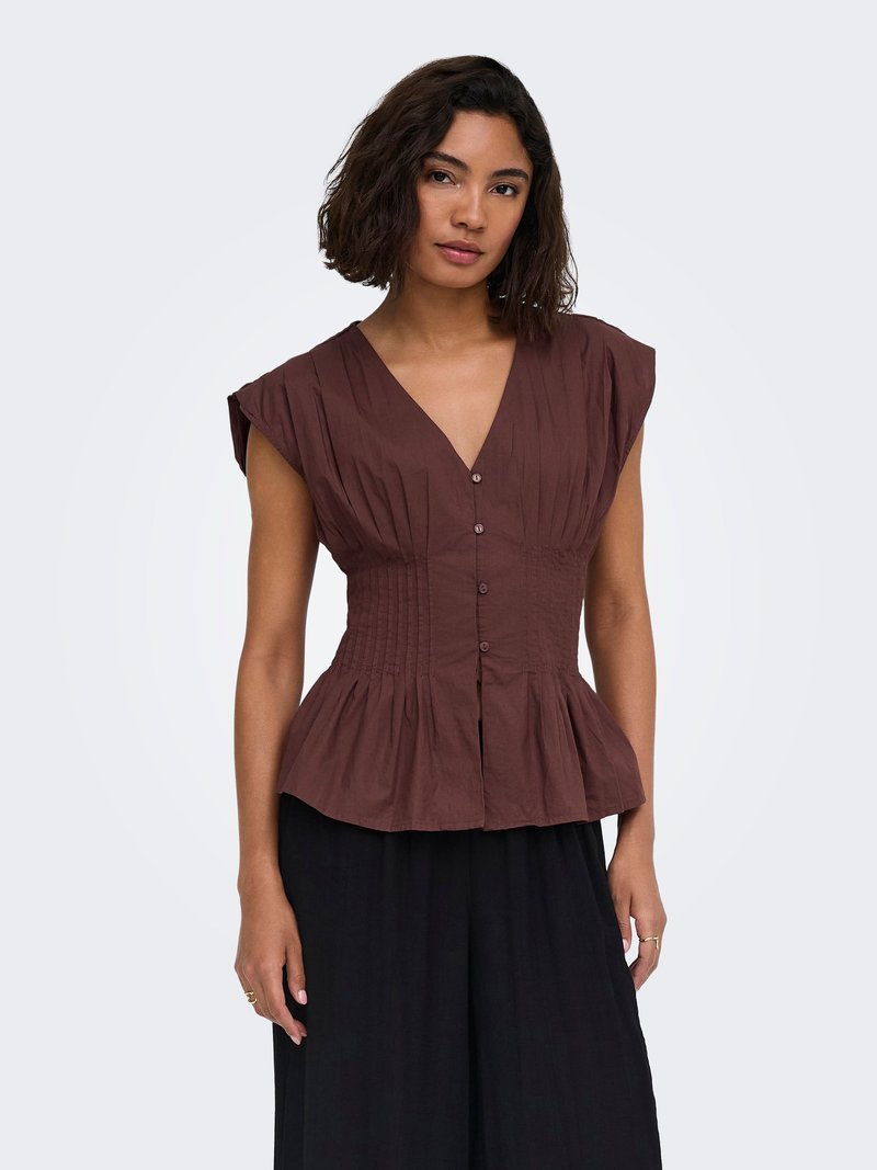 Mujer con cabello oscuro hasta los hombros que lleva una blusa marrón sin mangas con botones, cintura ajustada y pantalones negros de pierna ancha.
