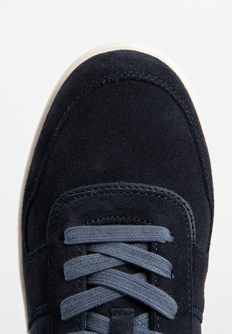 Marineblauwe suède sneaker met ronde neus, donkerblauwe veters en contrasterende witte rubberen zool. Gladde textuur met subtiele stiksels.