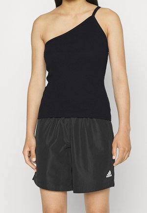 Kobieta ubrana w czarny top na jedno ramię oraz czarne spodenki Adidas z białym logo na lewej nodze, stojąca na białym tle.