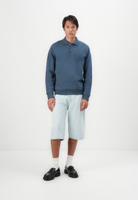 Marineblå genser med krage og knapper, kombinert med lyseblå denimshorts. Modellen har på seg svarte loafers og hvite ankelsokker.