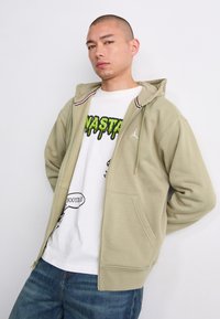 Zippered khaki hoodie with ribbed collar trim, white graphic T-shirt featuring colorful print, and denim jeans.