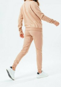 Krémová sportovní souprava skládající se z dlouhého rukávu a zúžených kalhot, vyrobená z měkké tkaniny s žebrovanými manžetami a pasem. Bílé tenisky.