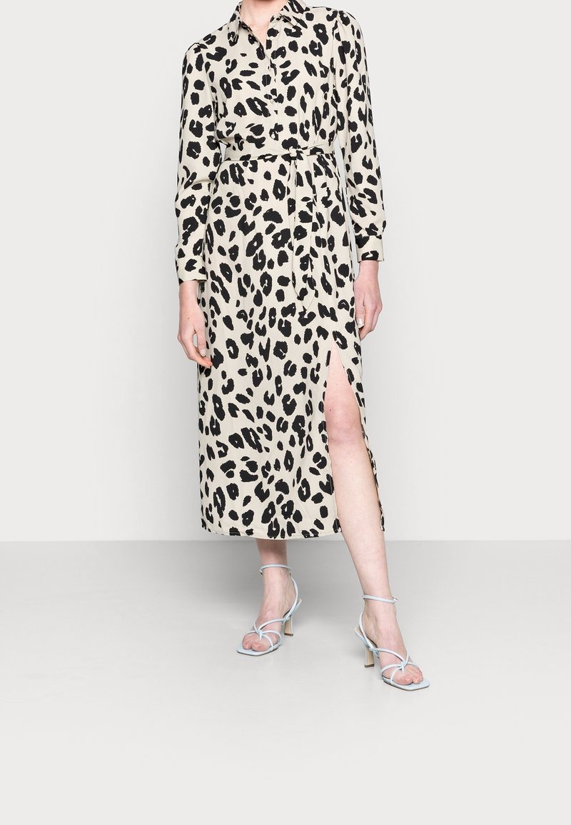 Femme portant une robe chemise midi beige à taches léopard noires, ceinture à la taille, manches longues, fente à la cuisse, et talons blancs à lanières.