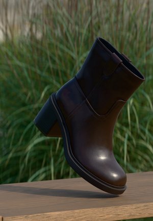 Stivaletto alla caviglia in pelle marrone scuro con tacco a blocco appoggiato su una superficie in legno, erba alta verde sullo sfondo sfocato.