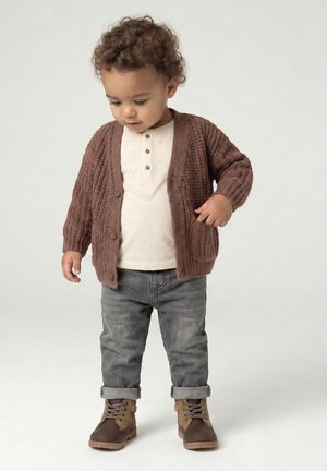 Toddler with curly hair wearing brown knit cardigan, white henley shirt, gray cuffed pants, and brown boots standing against a plain background.