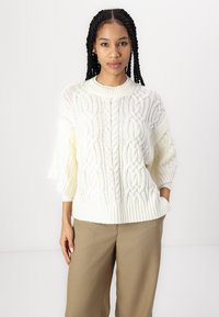 Maglione bianco a trecce con scollo rotondo e maniche a 3/4, con polsini e orlo a coste. Abbinato a pantaloni beige.
