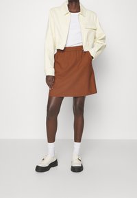 Giacca gialla cropped, t-shirt bianca, gonna A-line marrone, calzini bianchi e scarpe chunky bianche con suole nere, in piedi su uno sfondo neutro.